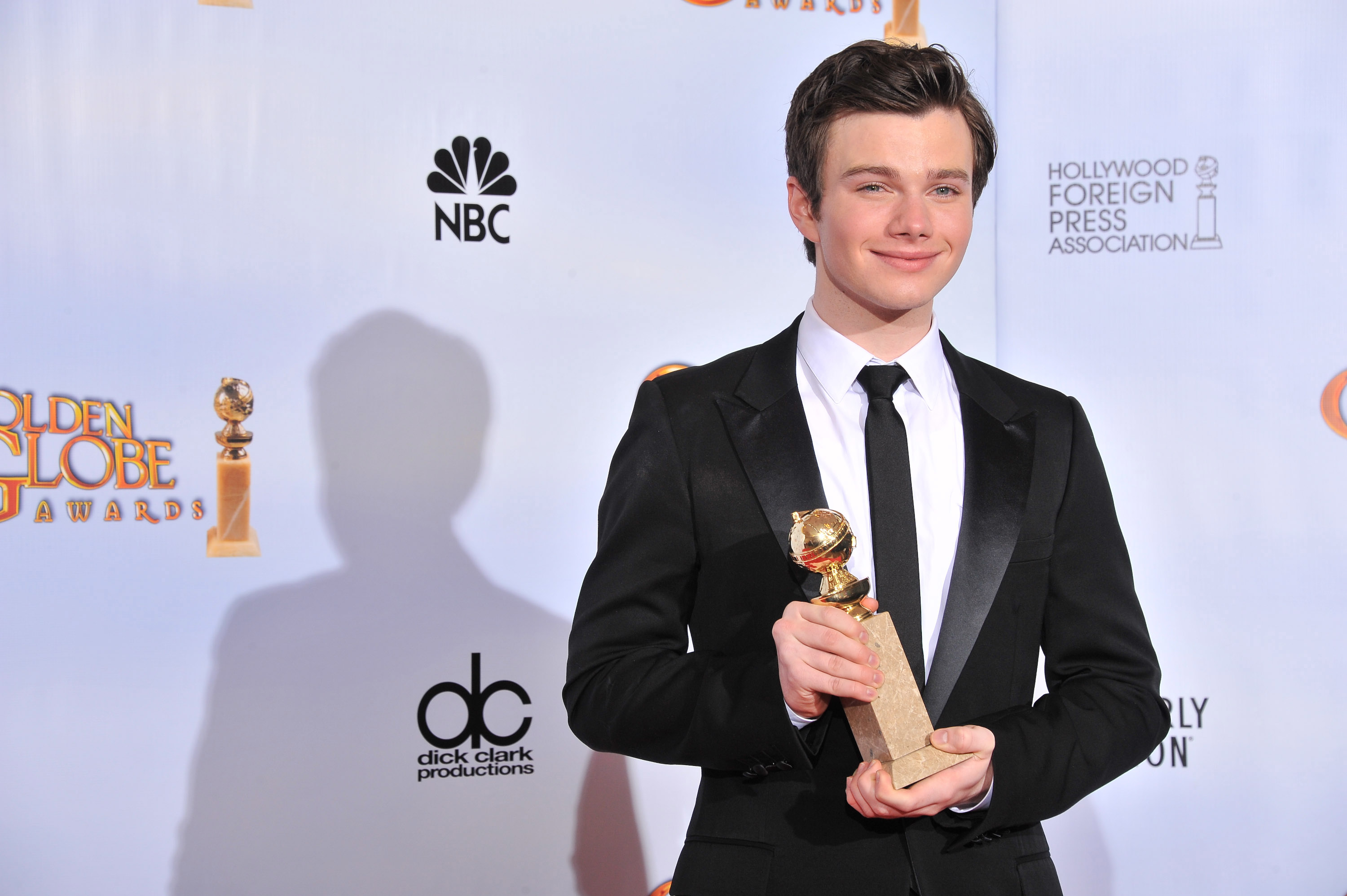 Chris Colfer on the red carpet holding a Golden Globe award, wearing a black suit with a black tie