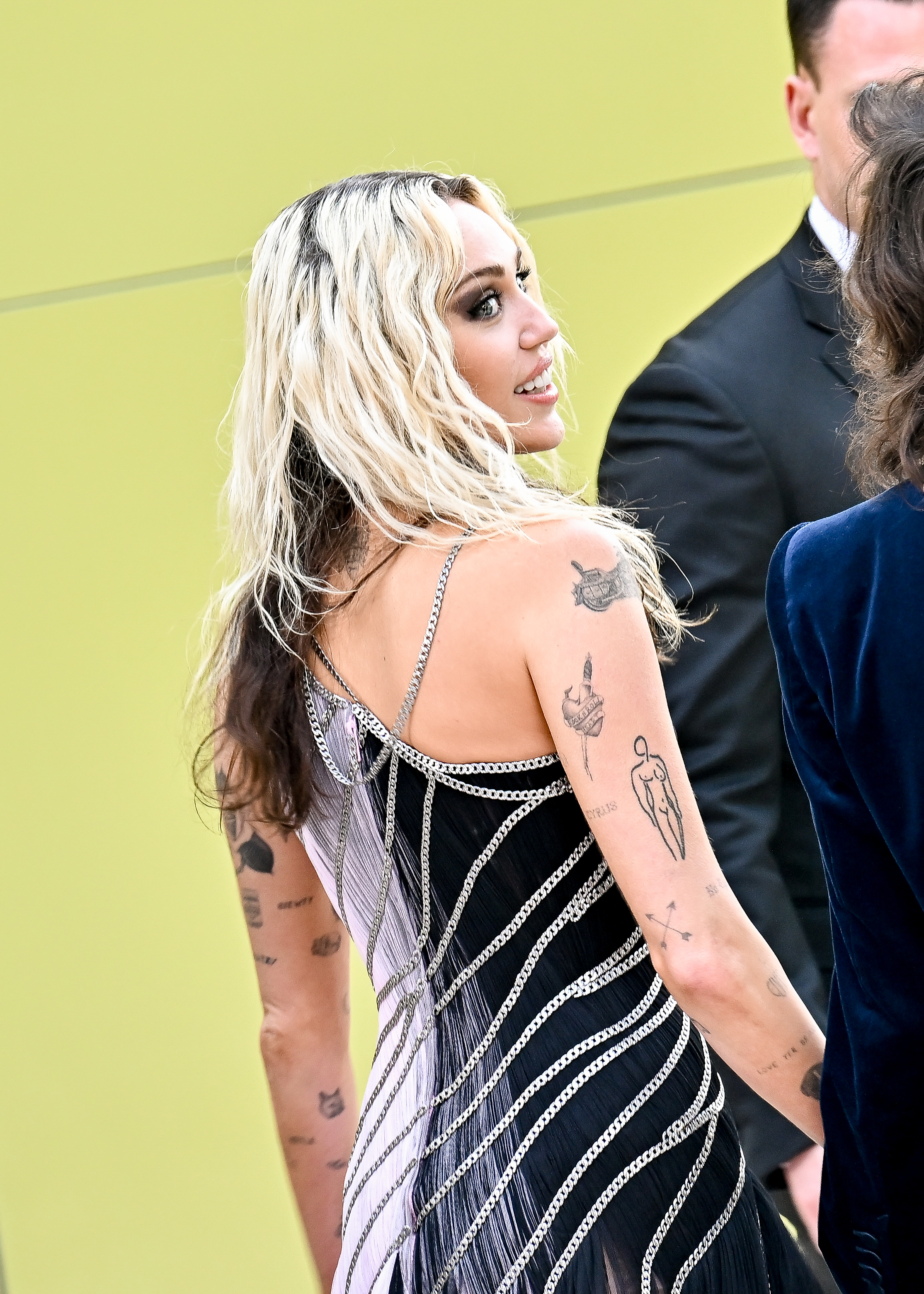 Miley Cyrus wearing a beaded, sleeveless dress with a zigzag pattern, smiling and looking over her shoulder at an event