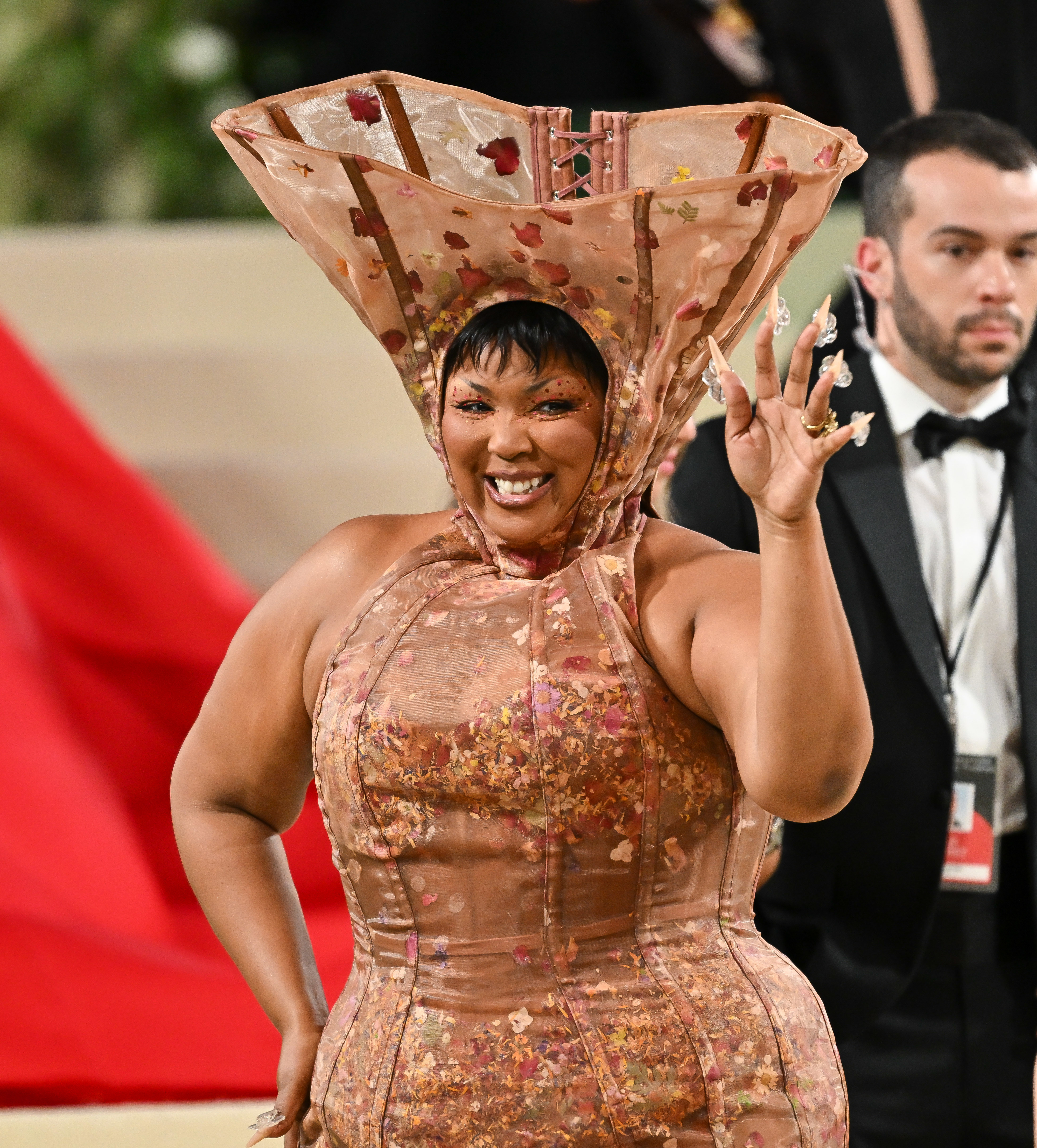 Lizzo smiles and waves on the red carpet, wearing an avant-garde dress with a unique, structured headpiece