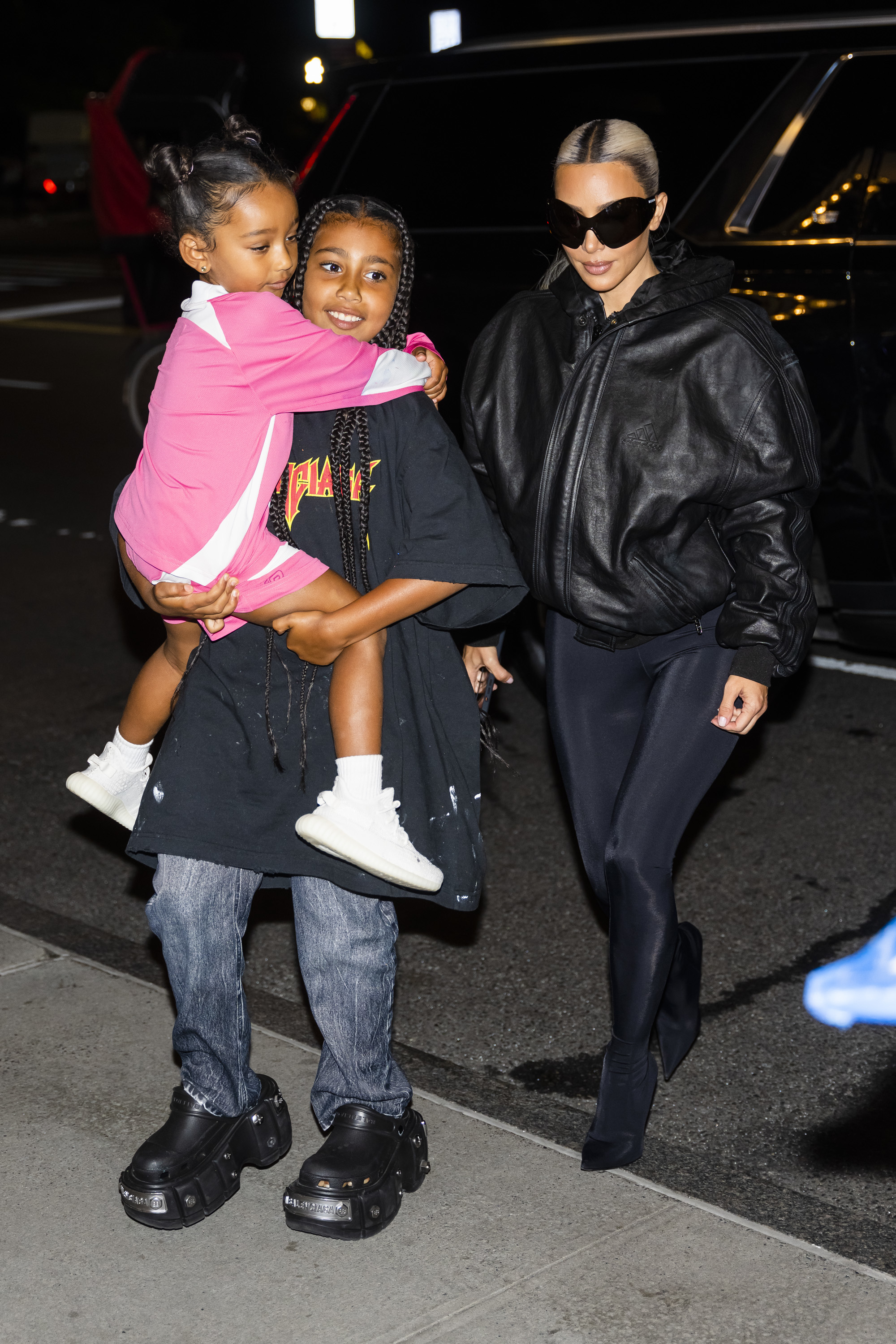 Kim Kardashian walks with her two children. One child is carrying the other, who wears a pink and white outfit