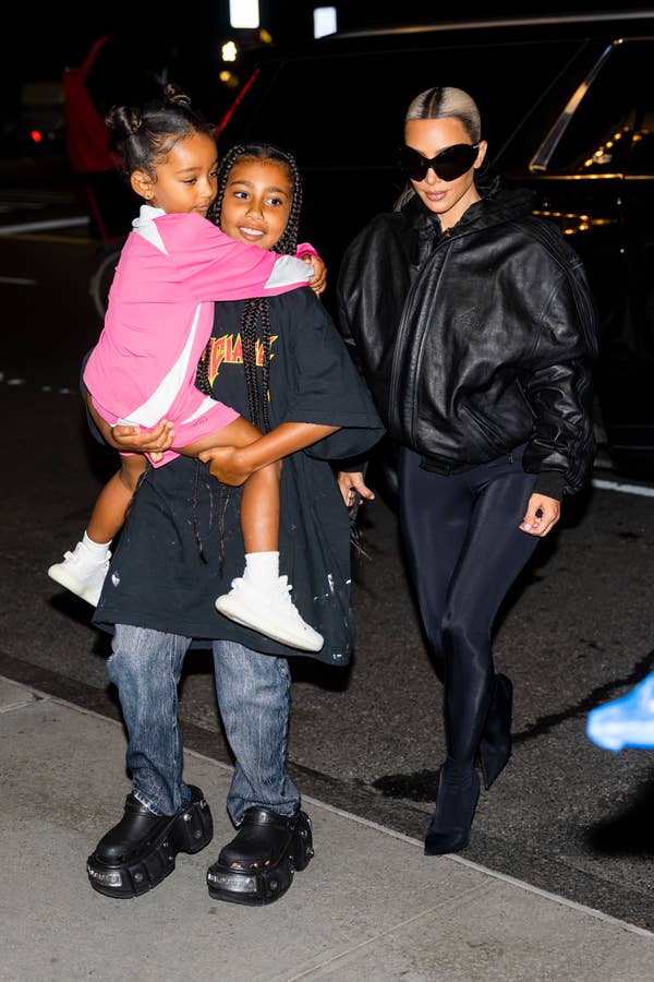 Kim Kardashian walks with her two children. One child is carrying the other, who wears a pink and white outfit