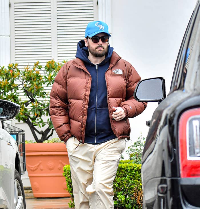 Scott wearing a cap, sunglasses, puffer jacket, and light pants walks beside a parked car