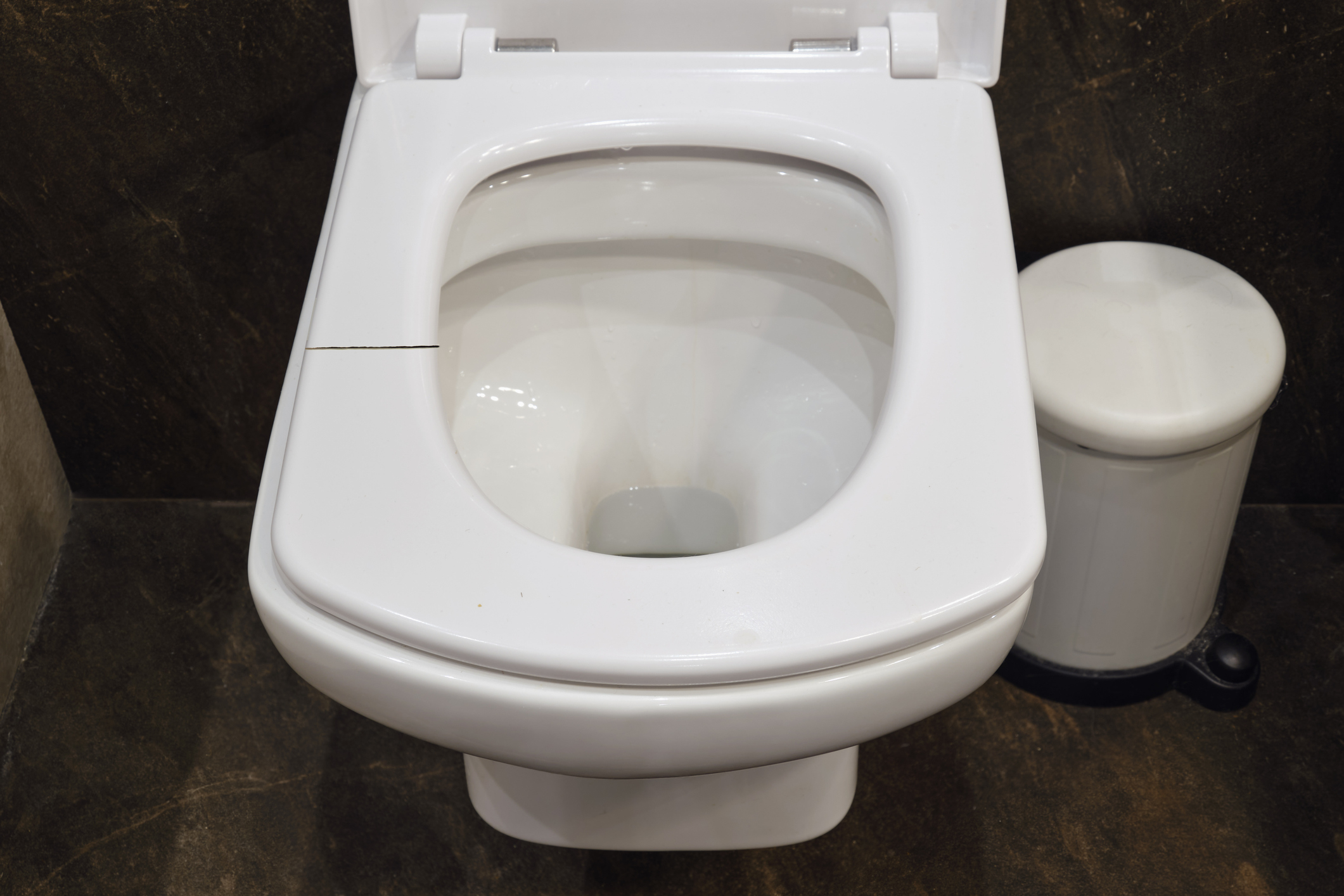 A white toilet with a visible crack on the seat, next to a white cylindrical trash can
