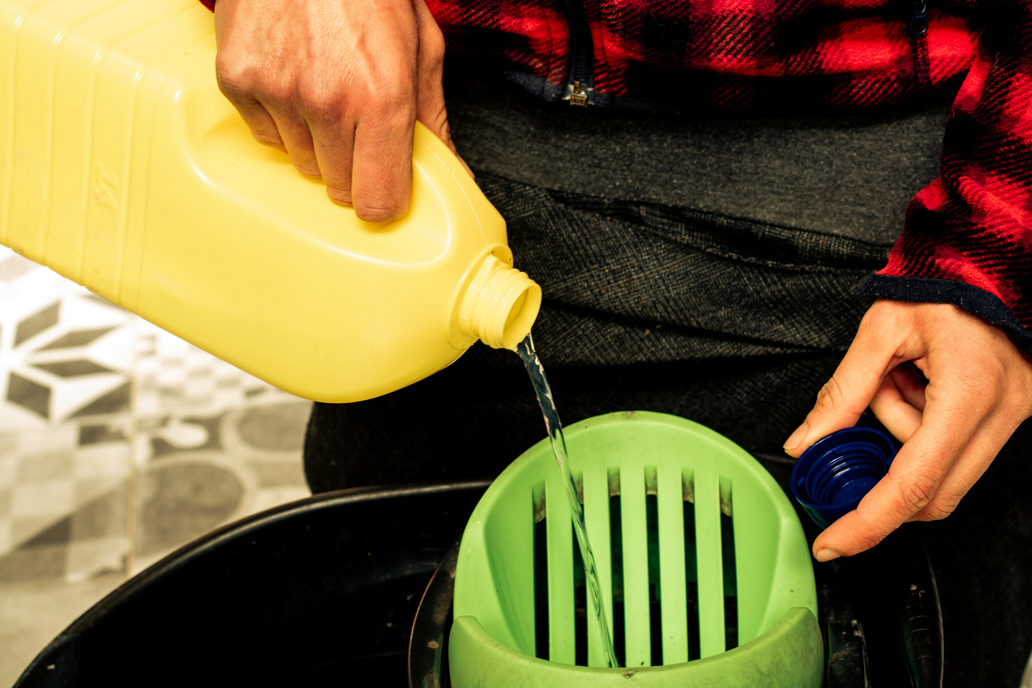 Person pouring liquid from a yellow container into a green machine. Only hands and part of the torso are visible