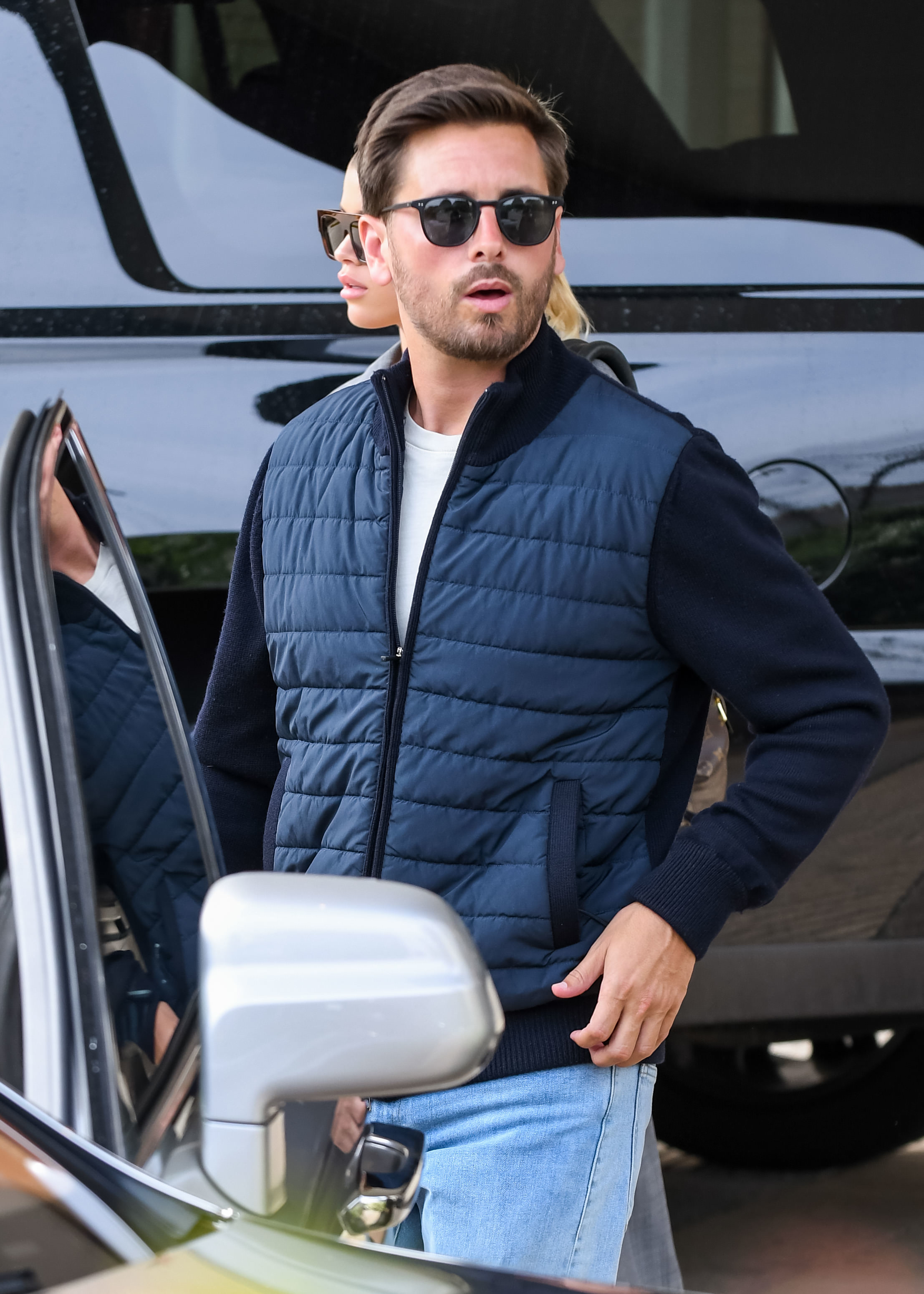 Scott Disick steps out of a car wearing a quilted vest, sweater, sunglasses, and light jeans