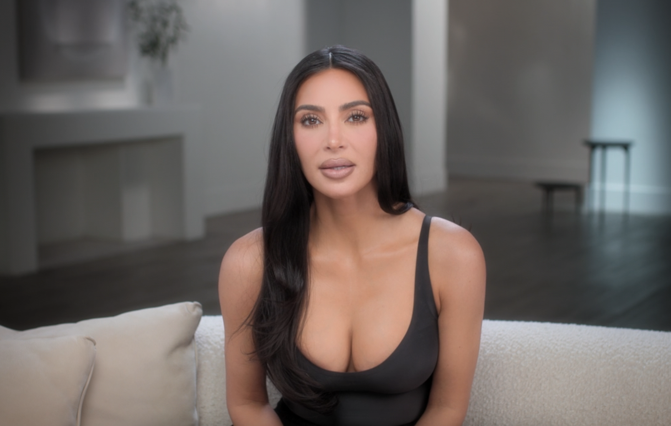 Kim Kardashian sits on a couch in a minimalist room, wearing a low-cut top