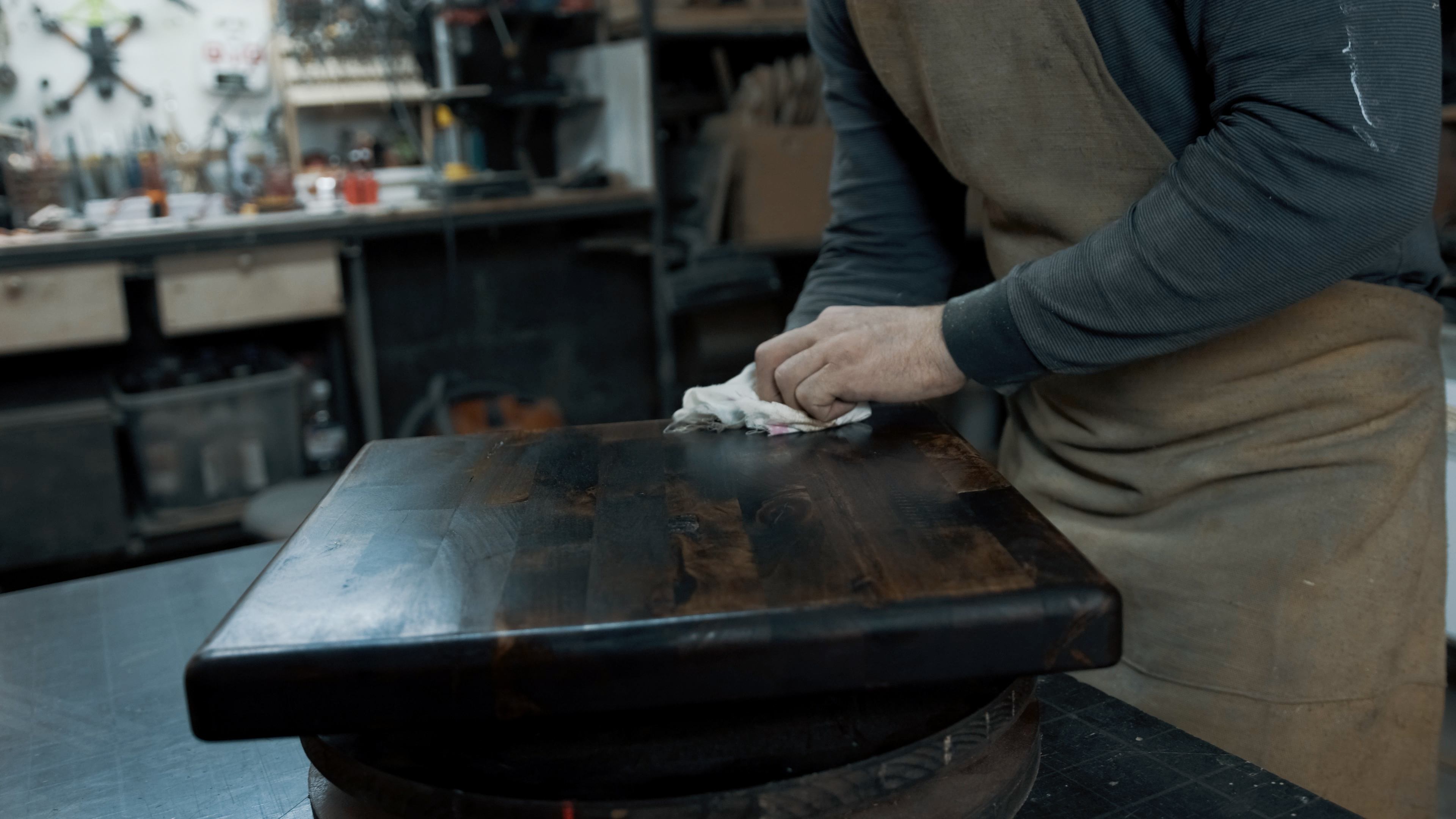 A person is sanding a wooden table in a workshop filled with tools and materials. Their face is not visible, and they are wearing an apron over a long-sleeve shirt