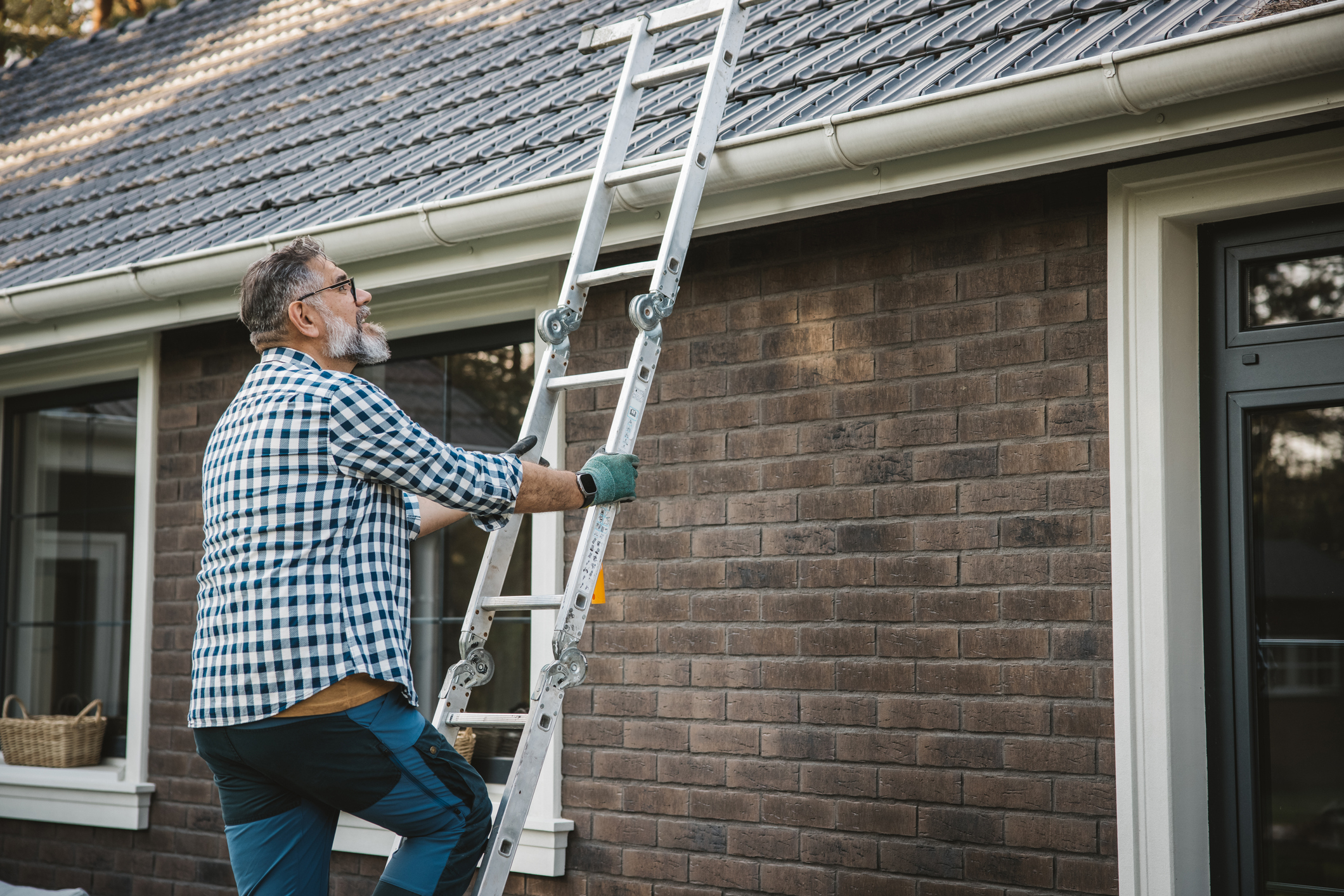 An older man in a plaid shirt and gloves climbs a ladder to clean the gutters on a brick house