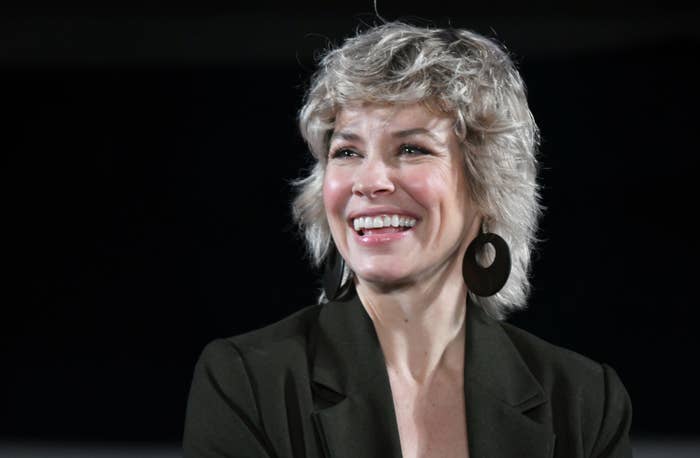 Lily, with short, wavy hair and large hoop earrings, wearing a dark blazer, smiles brightly