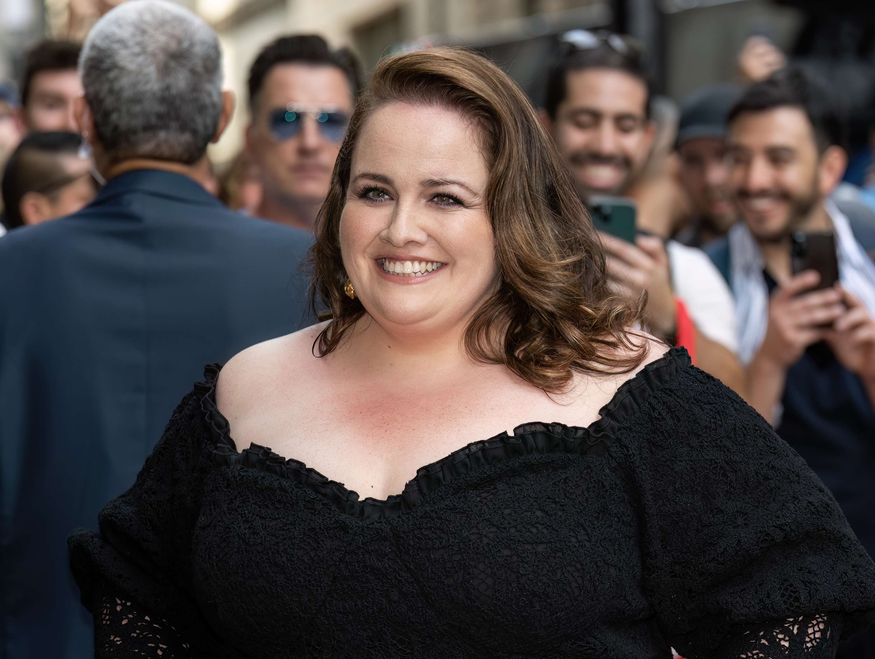 Jessica Gunning smiles at the camera, wearing an off-shoulder dress. People with phones and cameras can be seen in the background