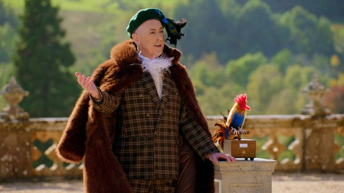 Alan Cumming, in a plaid suit and fur cape, stands beside a fake rooster on a pedestal. He is outdoors in a scenic setting
