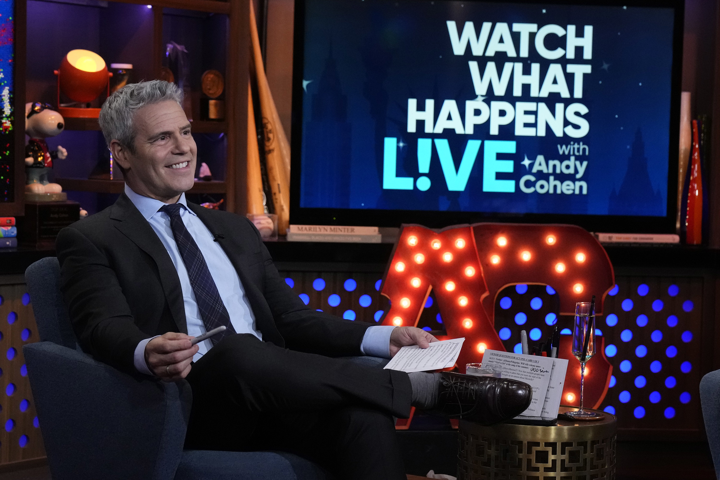 Andy Cohen sitting on his talk show set with the &quot;Watch What Happens Live with Andy Cohen&quot; logo in the background, holding cue cards and smiling