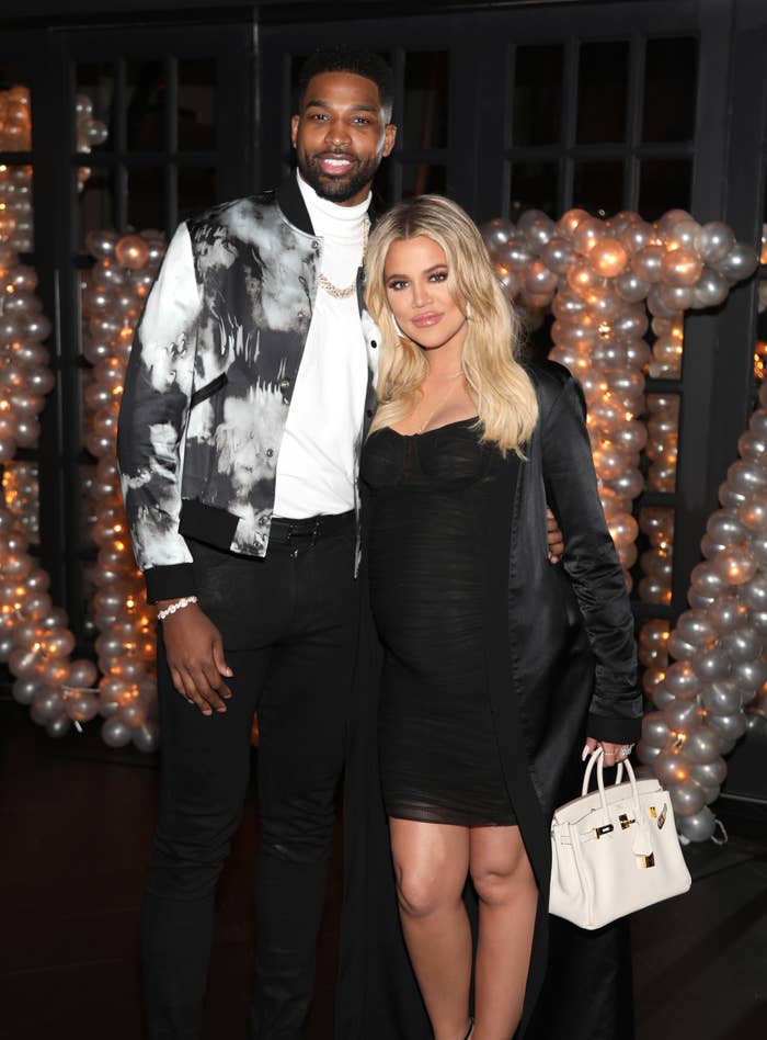 Tristan Thompson and Khloé Kardashian pose together; Tristan in a patterned jacket and Khloé in a short, dark dress and coat, carrying a white handbag