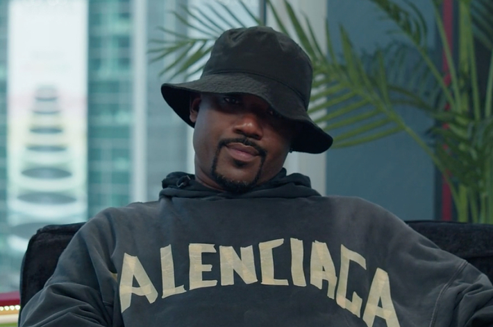 Ray J wearing a Balenciaga hoodie and a black bucket hat, seated indoors with a plant and buildings visible in the background