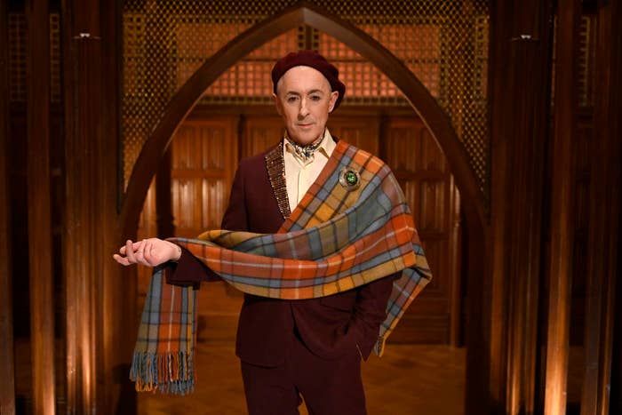 Alan Cumming poses in a stylish suit with an orange and blue plaid scarf, standing indoors with a wooden gothic arch backdrop