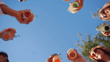 A group of people holding drinks up towards the sky, clinking glasses in a celebratory manner on a sunny day. Faces are mostly cropped out