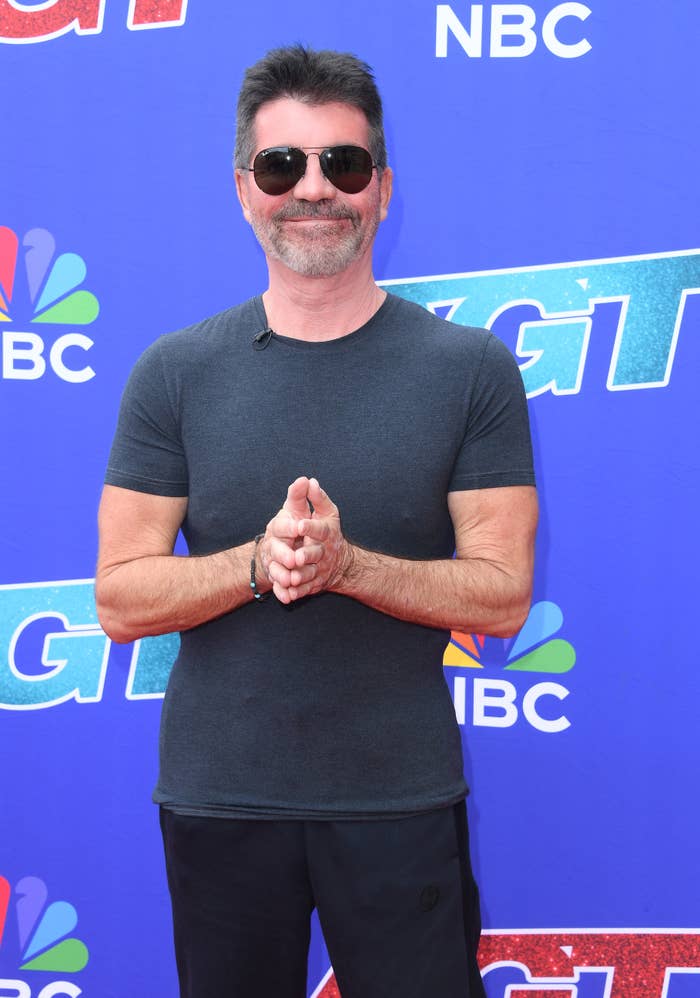 Simon Cowell on a red carpet for NBC's America's Got Talent, wearing a fitted dark T-shirt, dark pants, and sunglasses