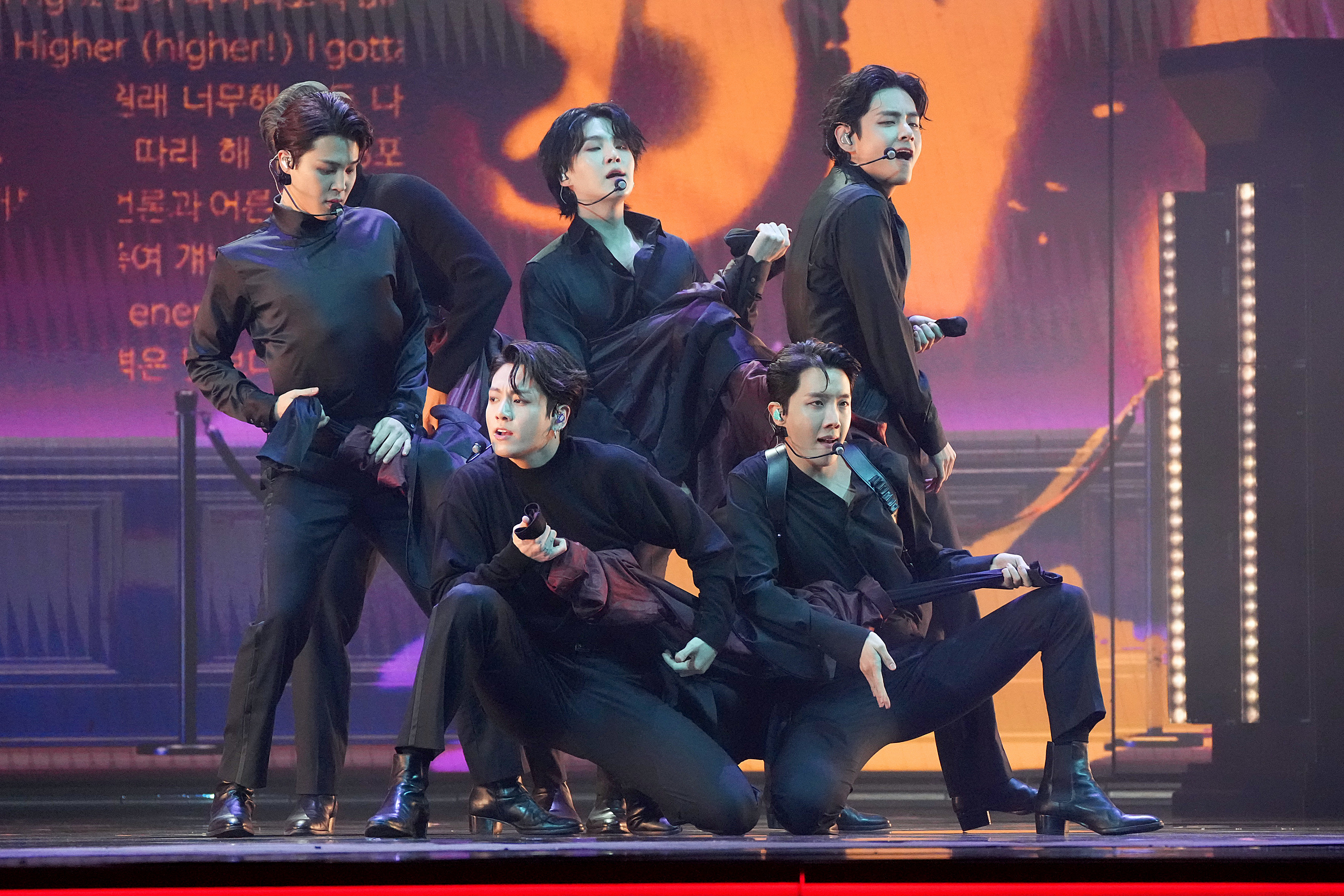 BTS members perform on stage in sophisticated black outfits, striking dynamic poses with microphones