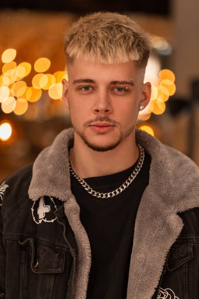 A man with short blond hair and a beard wears a faux fur-lined jacket, black t-shirt, and silver chain necklace. He has a piercing above his right eyebrow