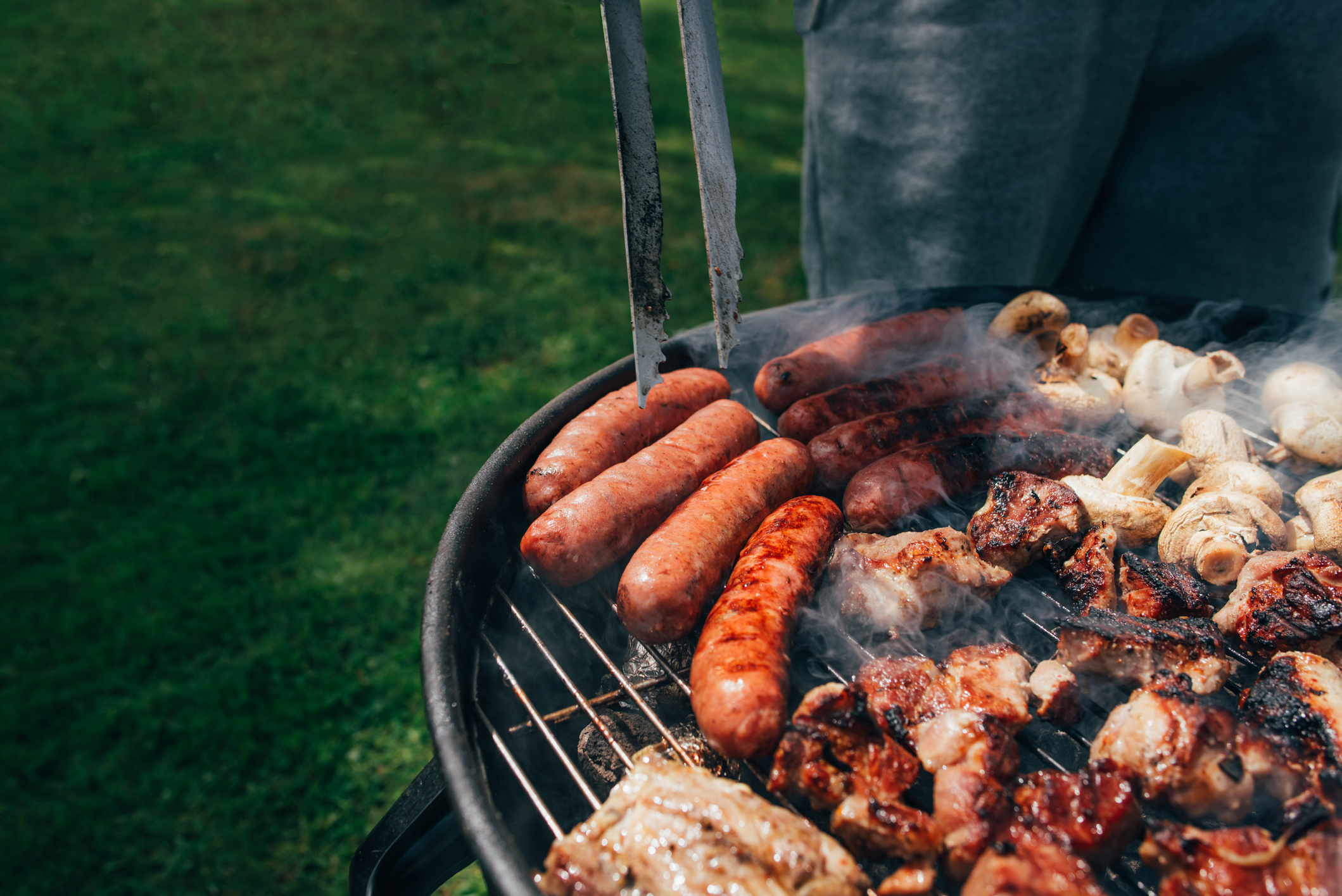 Close-up of a barbecue grill loaded with various meats, including sausages, kebabs, and mushrooms, with tongs holding one sausage