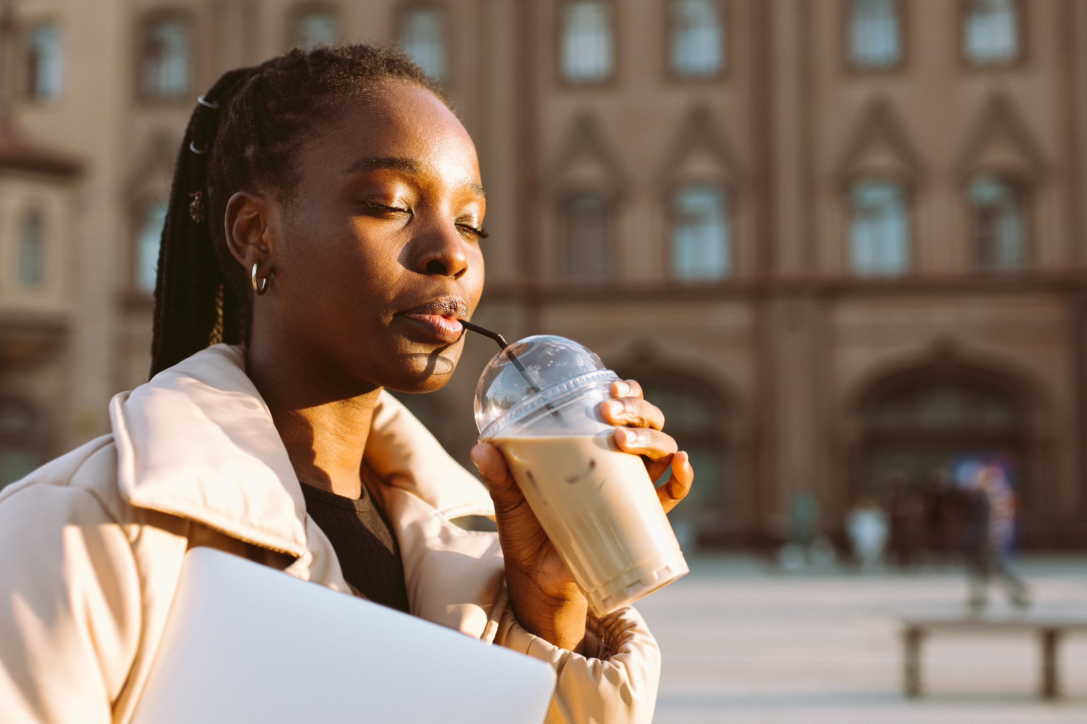 Person enjoying an iced coffee while holding a laptop outdoors in a city square