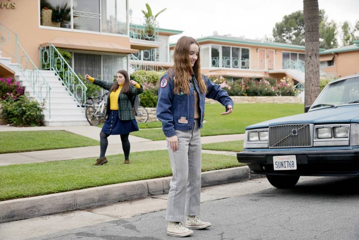 Kaitlyn Dever and Beanie Feldstein are pictured in a casual neighborhood scene from the movie &quot;Booksmart.&quot; Beanie dances in the background while Kaitlyn stands near a car