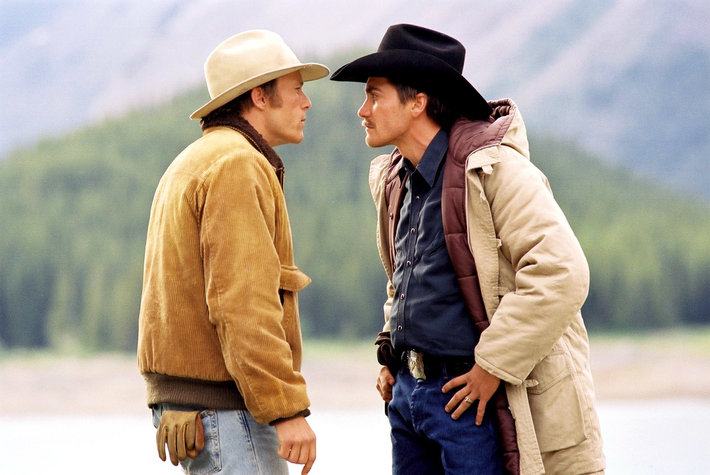 Heath Ledger and Jake Gyllenhaal stand facing each other in a tense scene from &quot;Brokeback Mountain,&quot; both wearing cowboy hats and layered jackets. Mountains are visible in the background