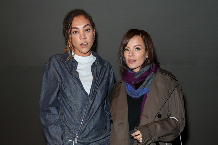 Two women, one in a denim suit holding a plaid purse, and the other in a trench coat with a fur bag, pose together against a plain backdrop. Names unknown