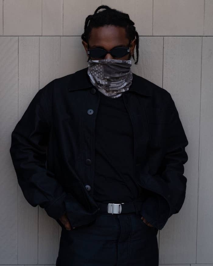 Musician Kendrick Lamar dressed in black attire, wearing a face covering and dark sunglasses, posing against a light wooden background