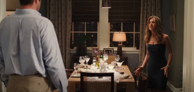 Jennifer Aniston, in a sleeveless dress, stands upset at a man in a button-up shirt and khaki pants. They are in a dining room with a table set for a meal