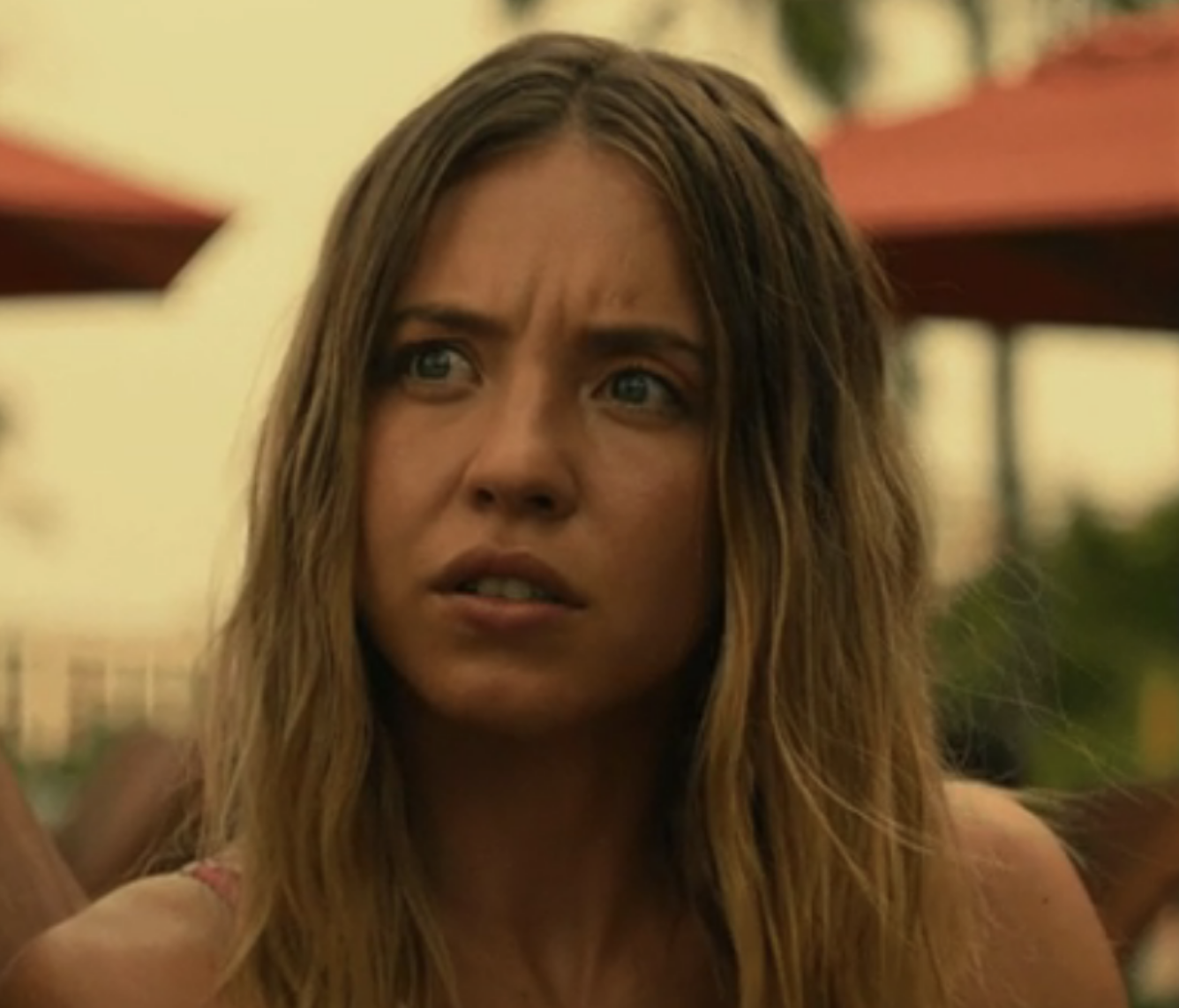 Sydney Sweeney with a worried expression. She has long, wavy hair and is outdoors with red umbrellas in the background