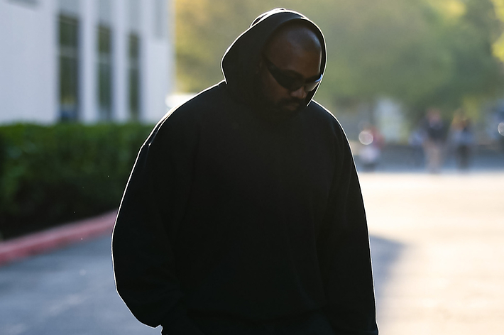 Ye is walking outdoors, wearing a black hoodie and sunglasses, with his head slightly bowed. The background is blurred, showing greenery and a pavement