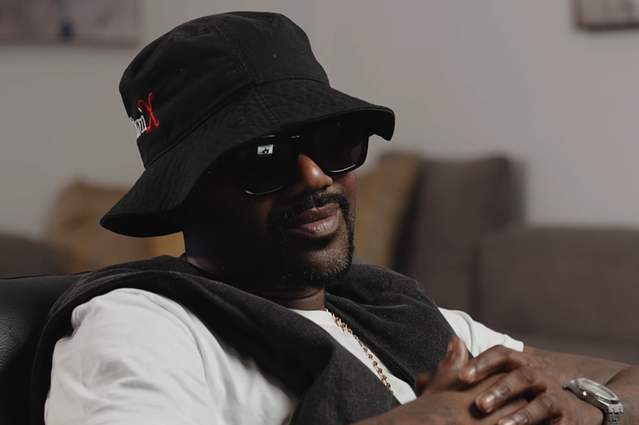 A man sits wearing a black bucket hat, dark sunglasses, and a white shirt with a dark jacket draped over his shoulders