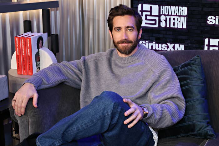 Jake Gyllenhaal wearing a casual grey sweater and jeans, seated on a couch in the SiriusXM studio during an interview on The Howard Stern Show