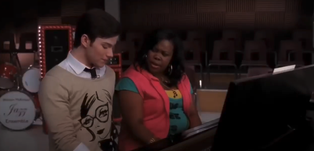 Chris Colfer and Amber Riley are singing at a piano. Chris is wearing a sweater with a face design, and Amber is wearing a vest over a colorful T-shirt