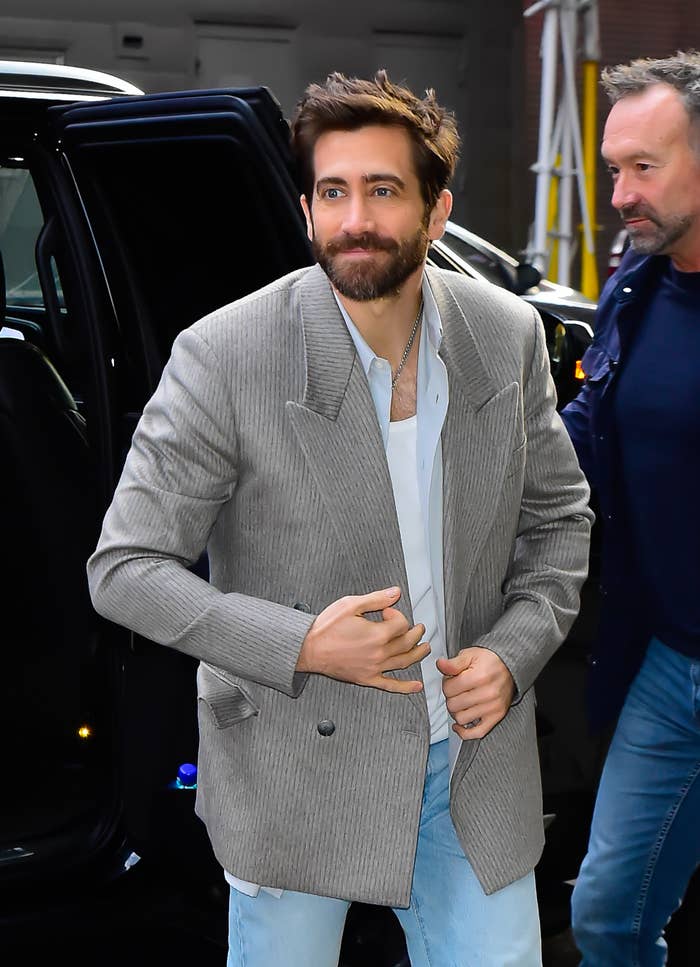 Jake Gyllenhaal smiling in a casual outfit with a blazer and jeans, standing next to a man outdoors