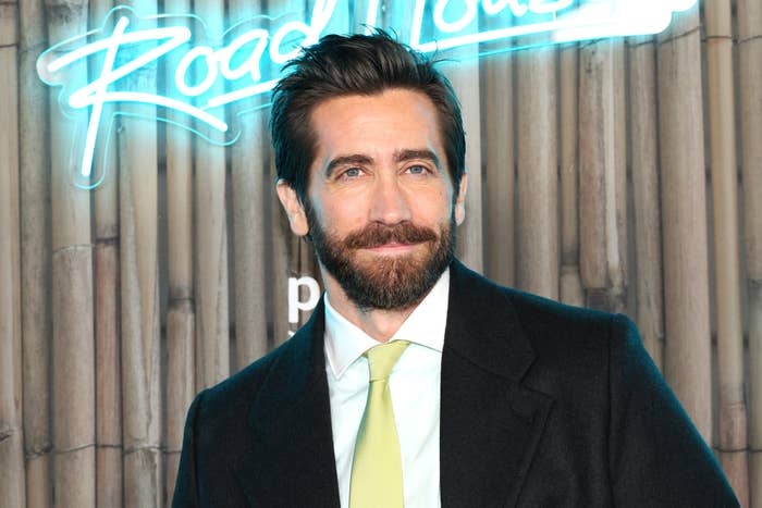 Jake Gyllenhaal at the "Road House" premiere, wearing a suit with a white shirt and a green tie. The background features the movie's neon title sign