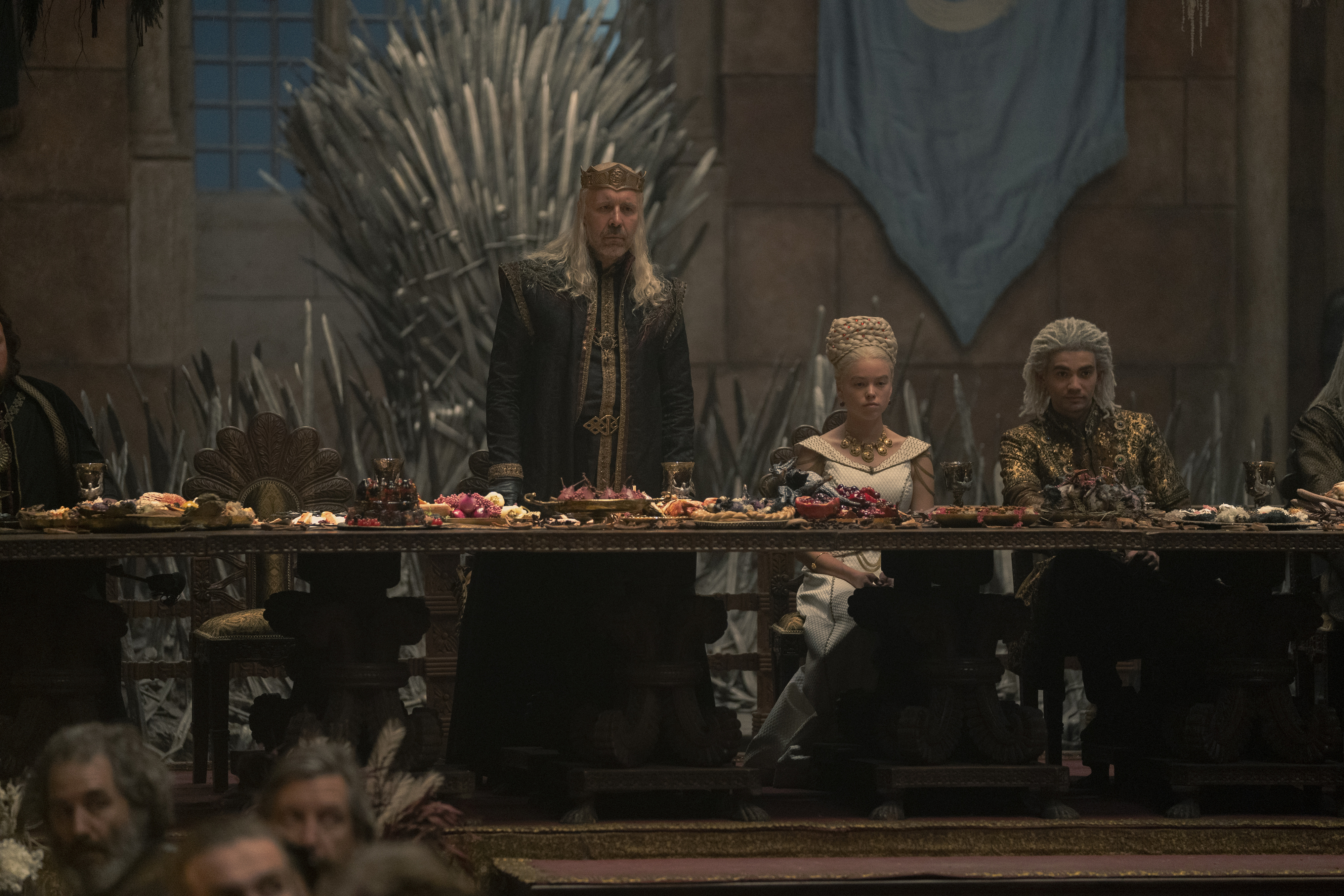 King Viserys Targaryen, Princess Rhaenyra Targaryen, and Ser Otto Hightower are seated at a lavish feast in a grand hall with the Iron Throne in the background