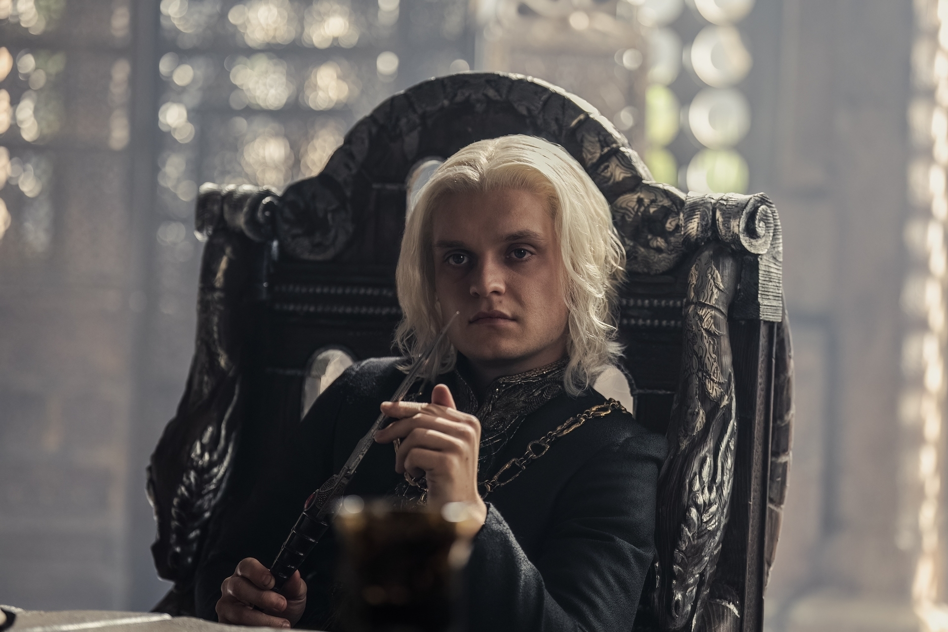 Tom Glynn-Carney as Aegon Targaryen sits on a grand chair, holding a dagger, in a medieval-style setting on a TV show