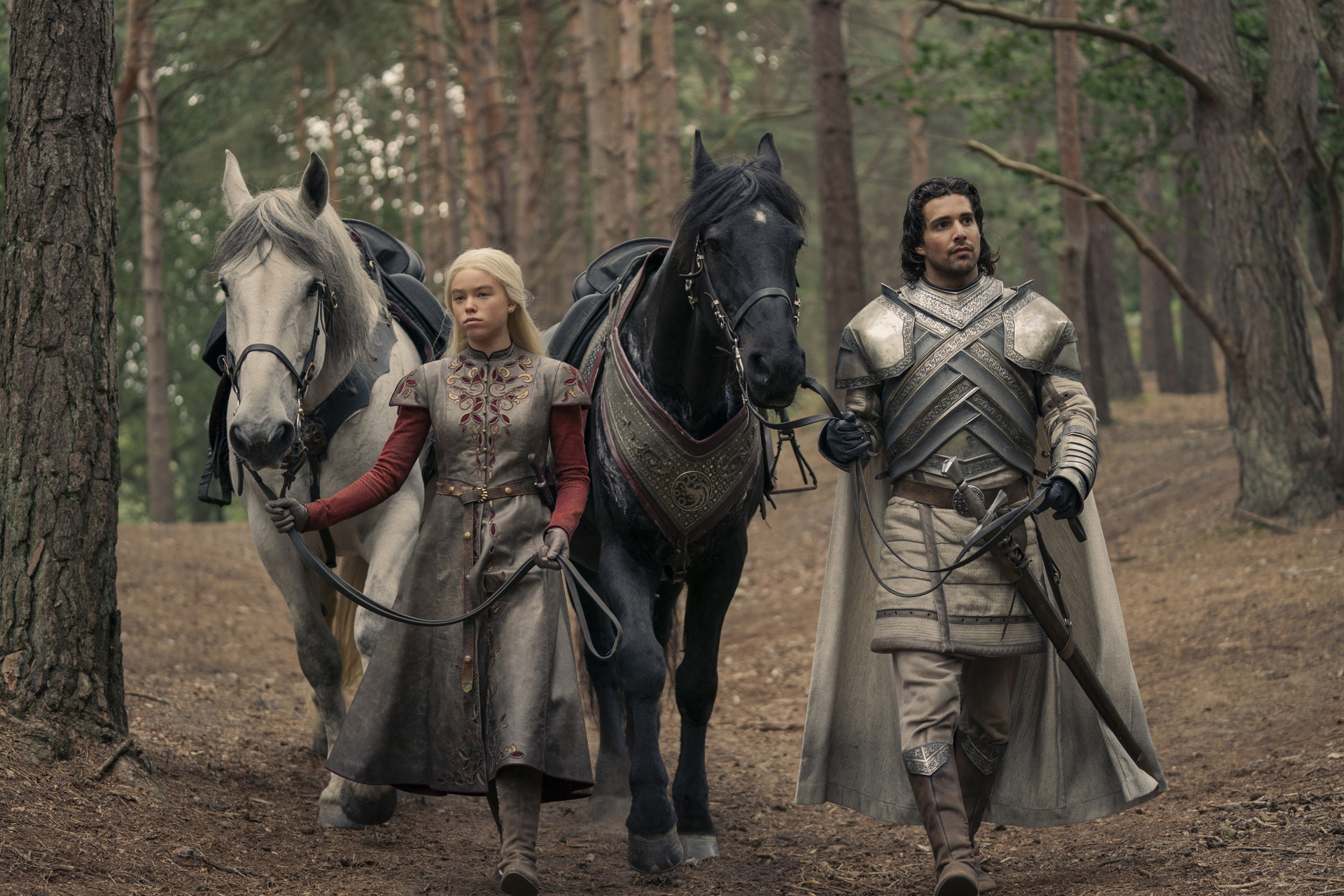 Emilia Clarke as Daenerys Targaryen walking with a white horse, and Michiel Huisman as Daario Naharis with a black horse, both dressed in medieval attire in a forest