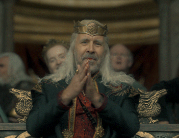 Paddy Considine as King Viserys I Targaryen, wearing a crown and medieval-style robes, claps while seated on a throne in a scene from &quot;House of the Dragon.&quot;