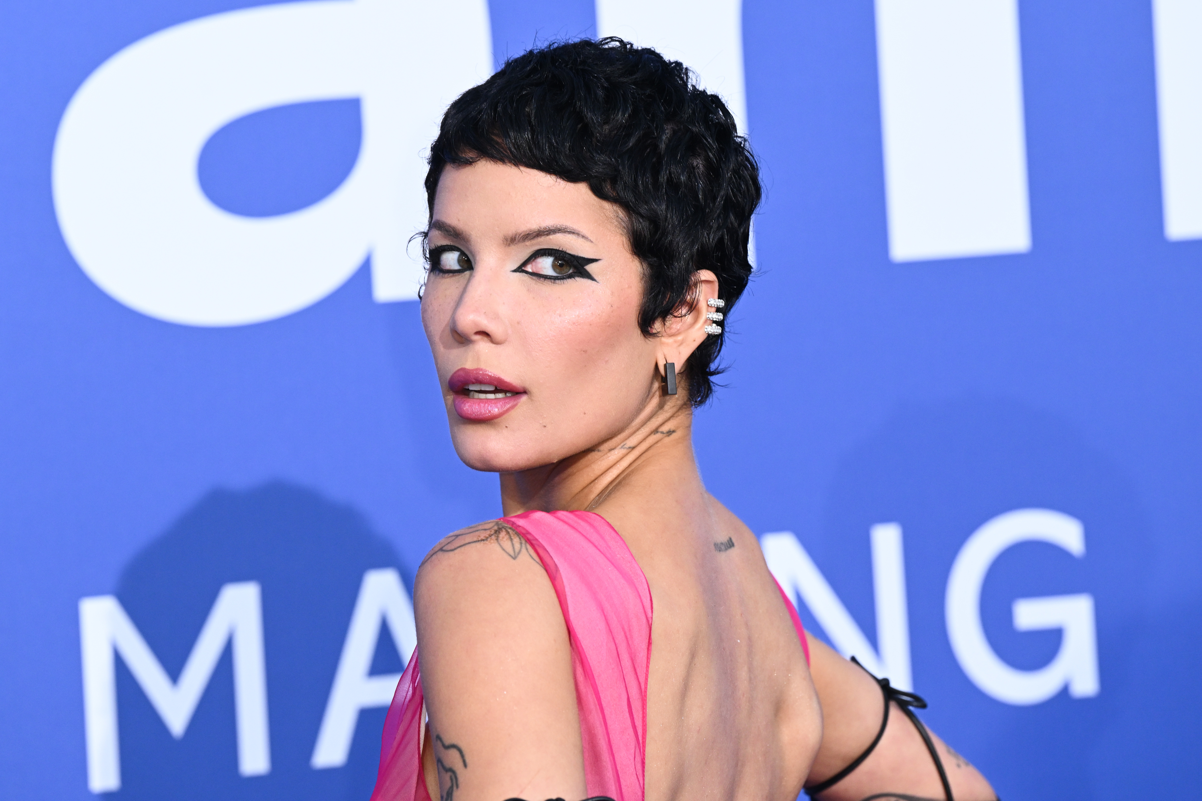Halsey poses on the red carpet, wearing a backless dress with unique eye makeup, a pixie haircut, and visible tattoos