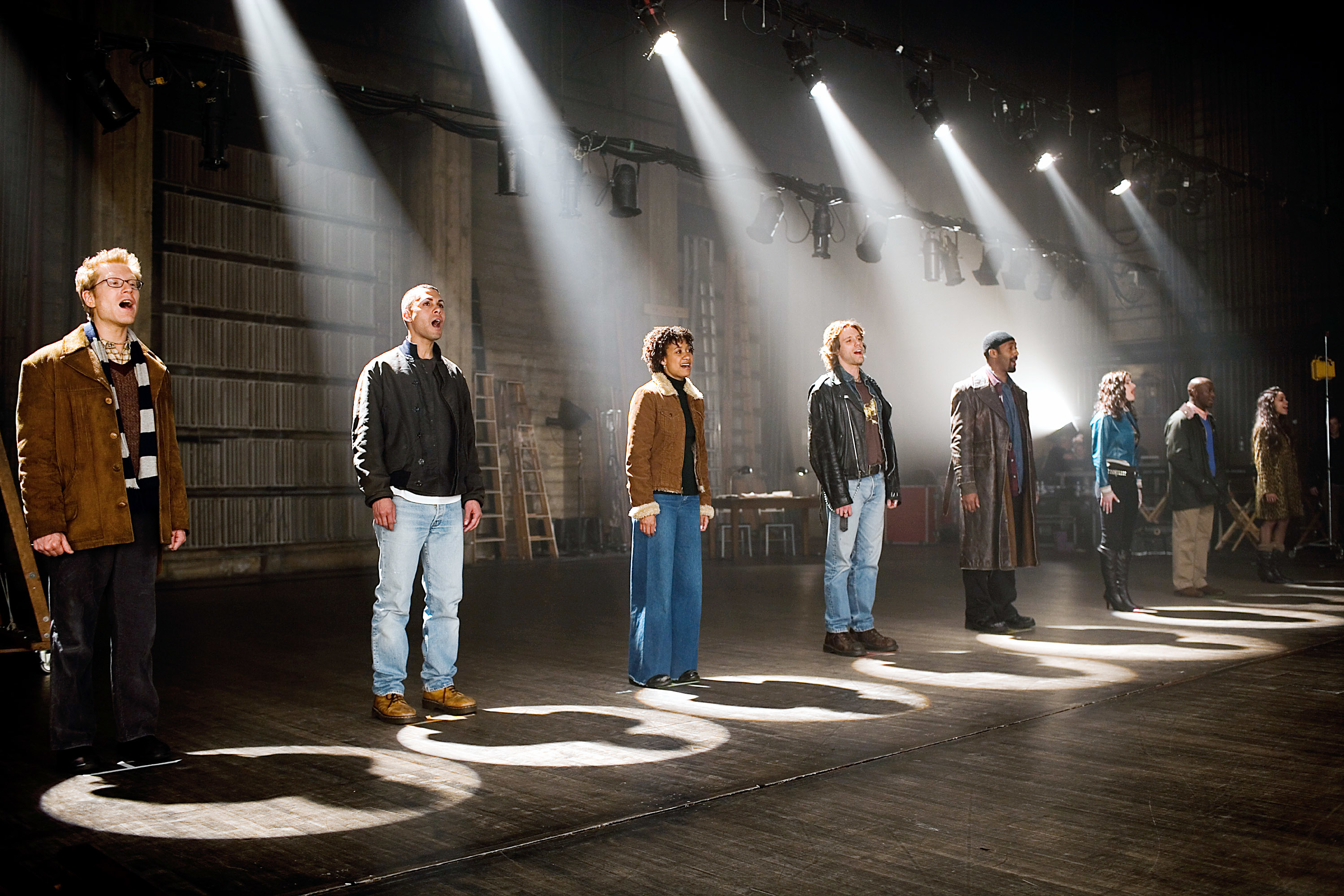 Actors in the 2005 film &quot;Rent&quot; stand in a line on stage, facing forward as a spotlight shines on each of them separately