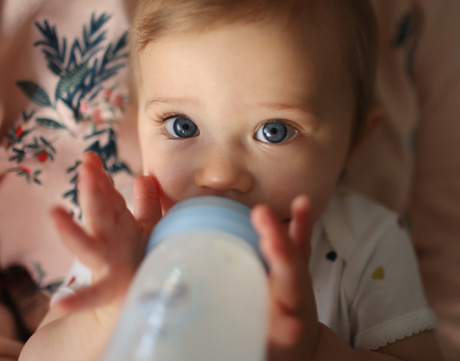 A baby with blue eyes holds a baby bottle with both hands and looks at the camera. There&#x27;s a patterned fabric in the background