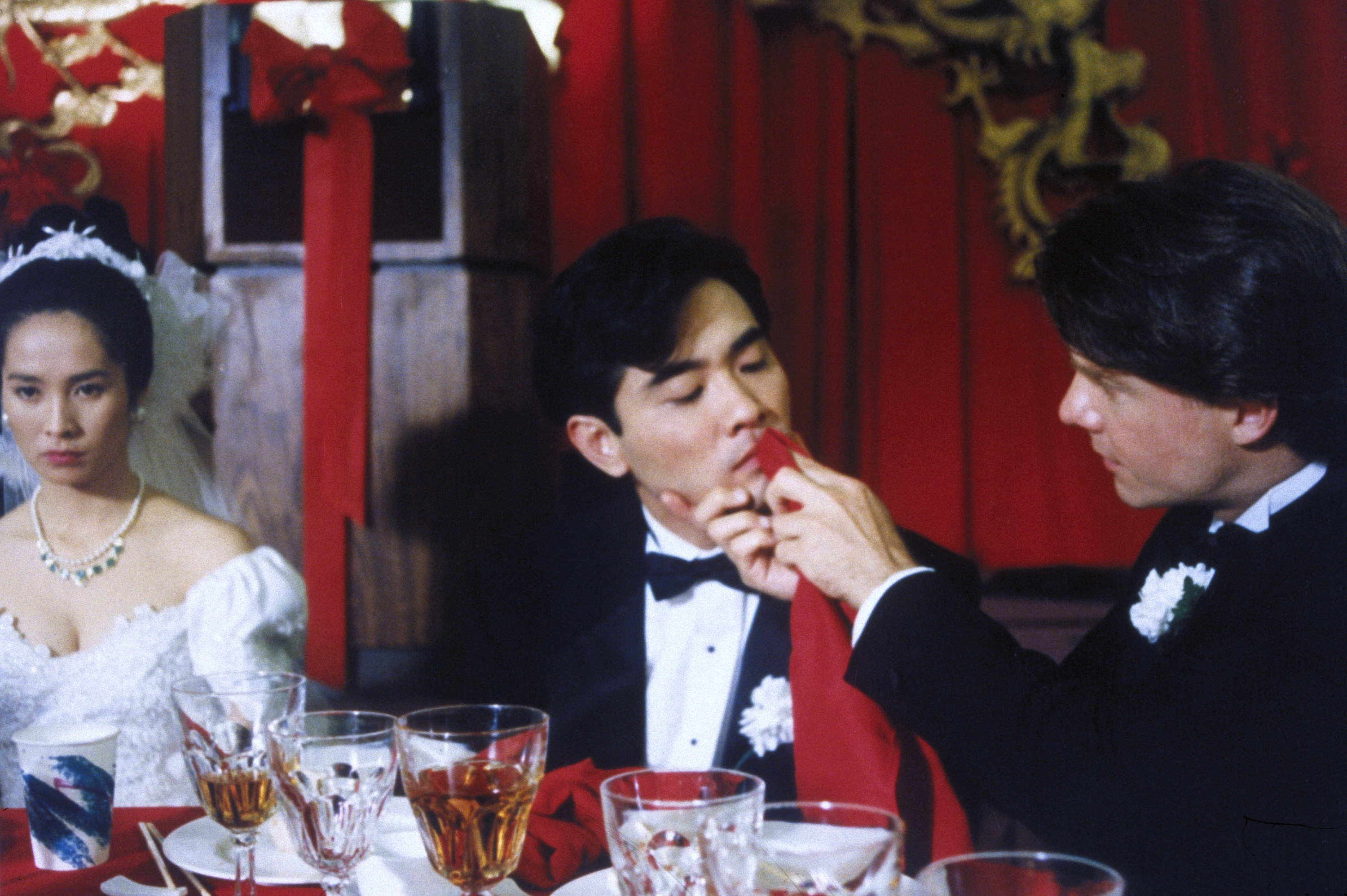 Actors in formal attire in a wedding scene from &quot;The Wedding Banquet,&quot; directed by Ang Lee. The groom is being fed by another man while the bride looks on