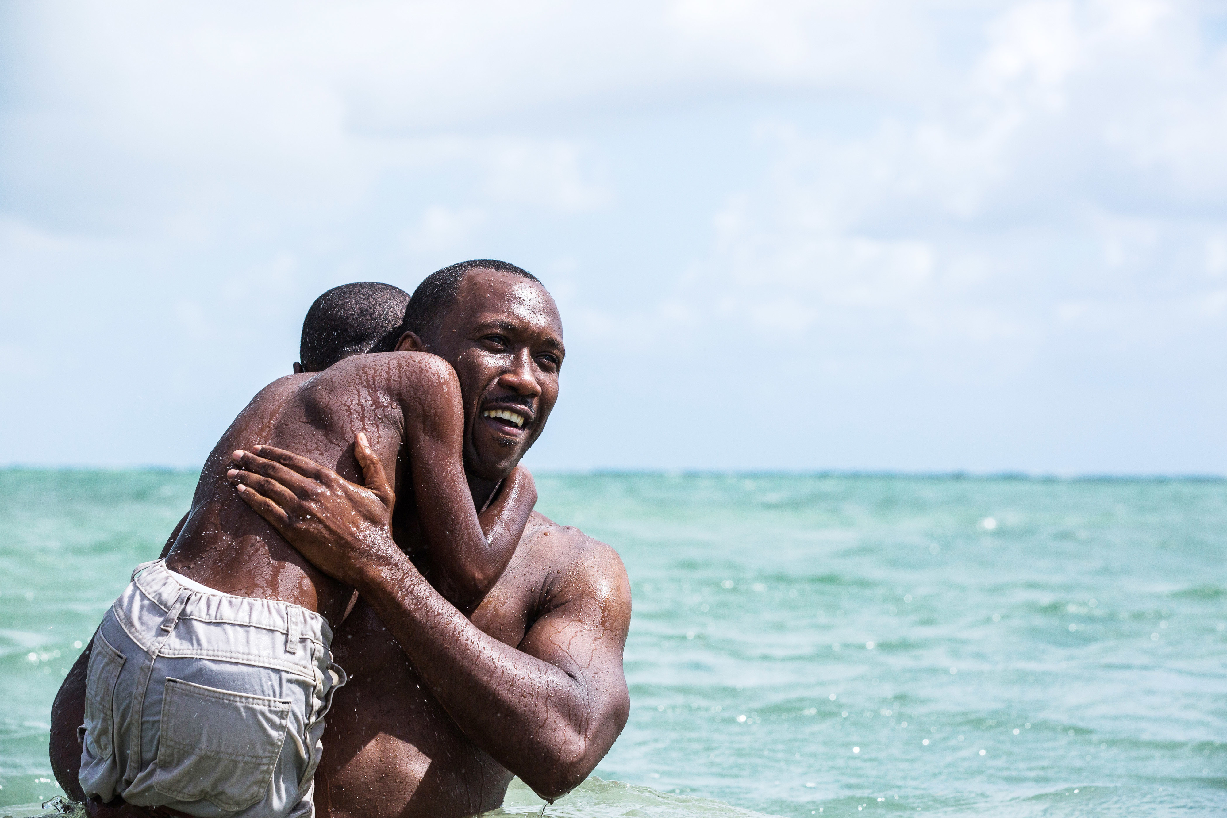 Mahershala Ali smiling and holding a young boy in his arms while standing in the ocean in a scene from &quot;Moonlight&quot;
