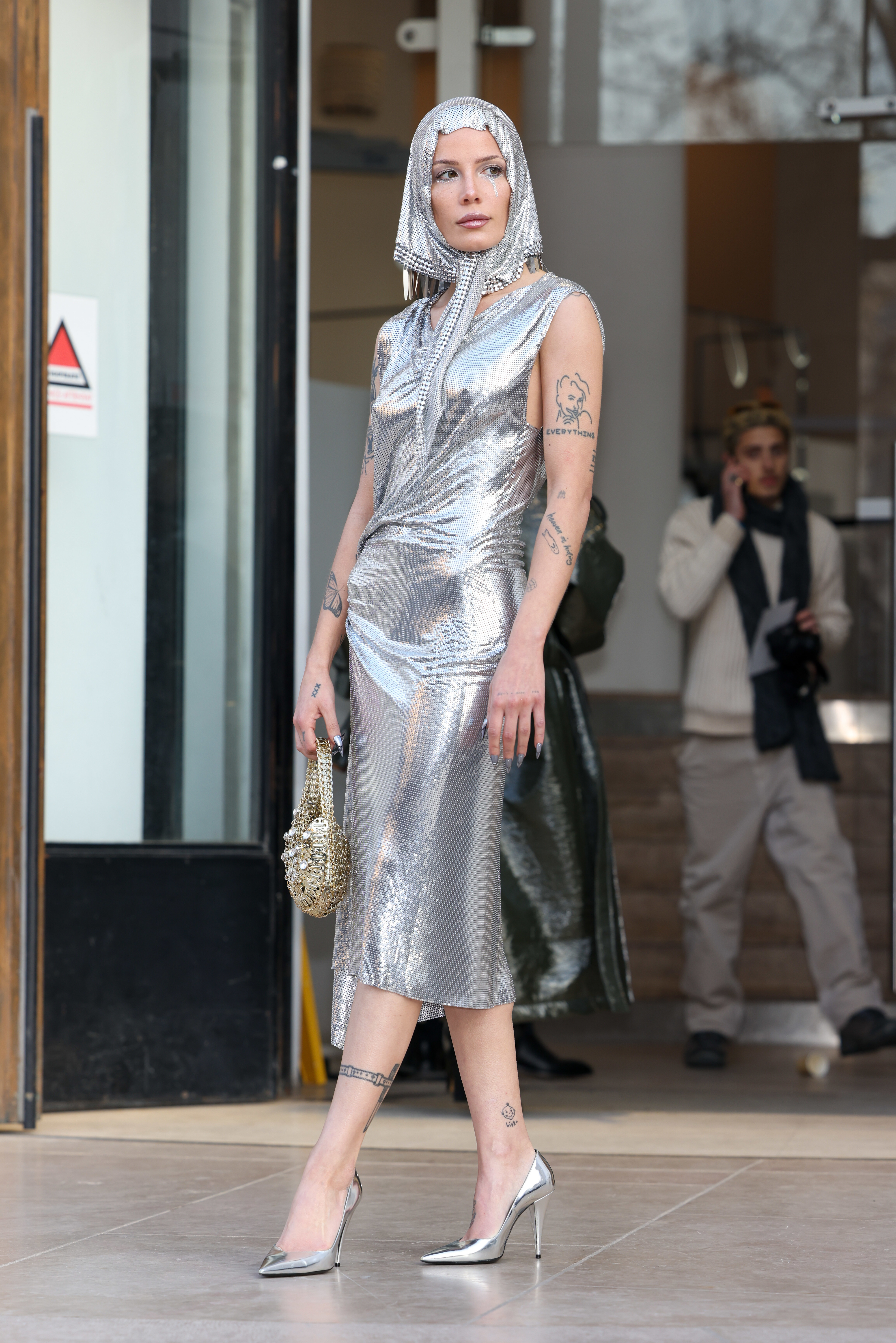 Halsey wearing a shimmering metallic dress and head covering, holding a small bag, and walking in front of a building