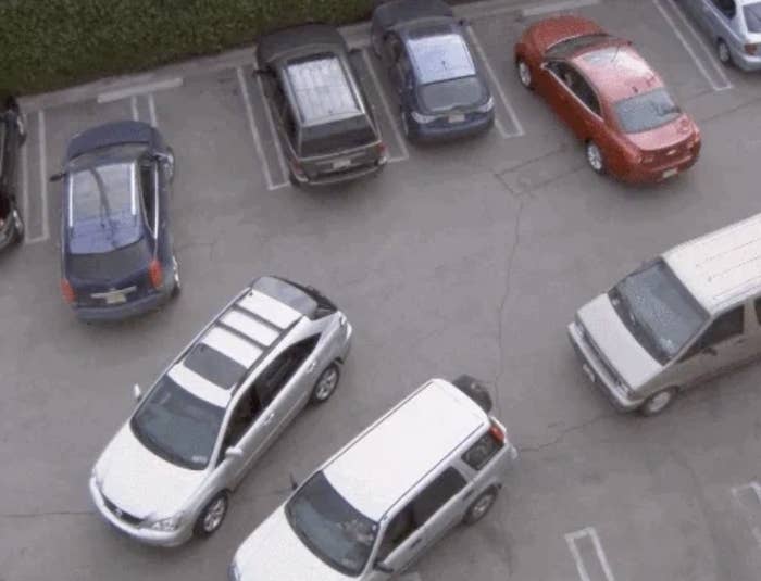 Cars are parked in a lot, with some vehicles improperly parked. Two cars are maneuvering to correct their positions. One car is driving into a spot