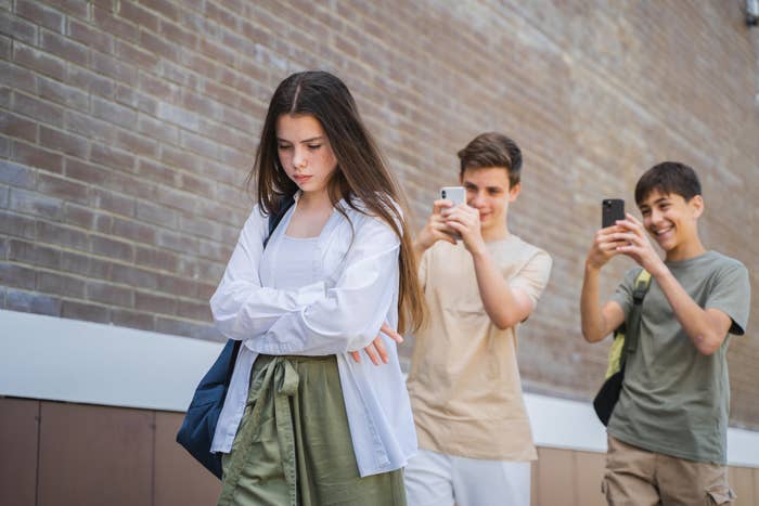 A girl walks with her arms crossed, looking down. Two boys behind her are taking photos with their phones and laughing