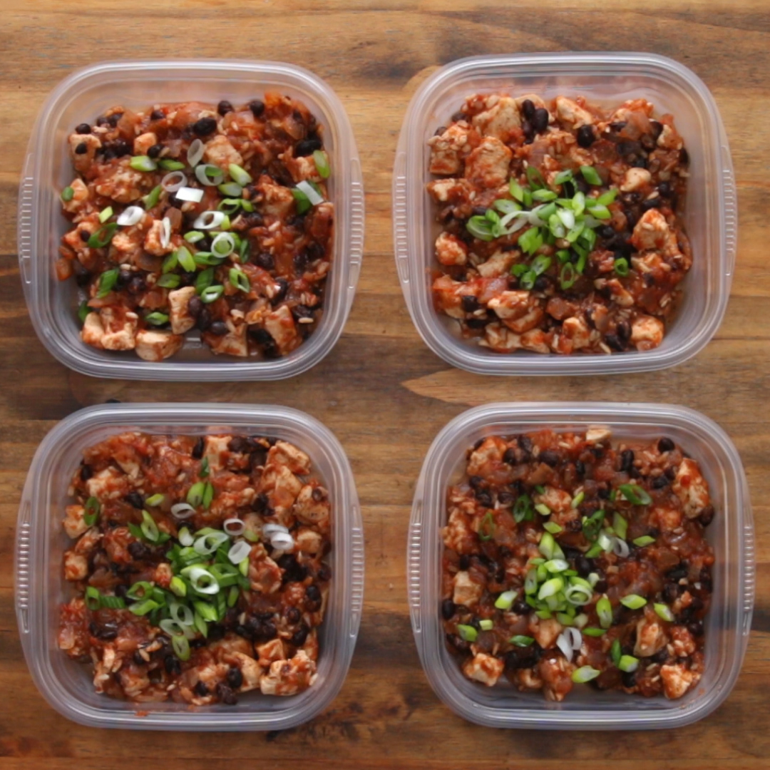 Four plastic containers filled with cooked chicken, black beans, and topped with chopped green onions are arranged on a wooden surface